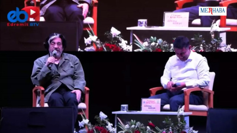 edremit'te konuşma yapan okan bayülgen'in tv de görünüsü: koltukta oturuyor ve soru cevaplıyor.